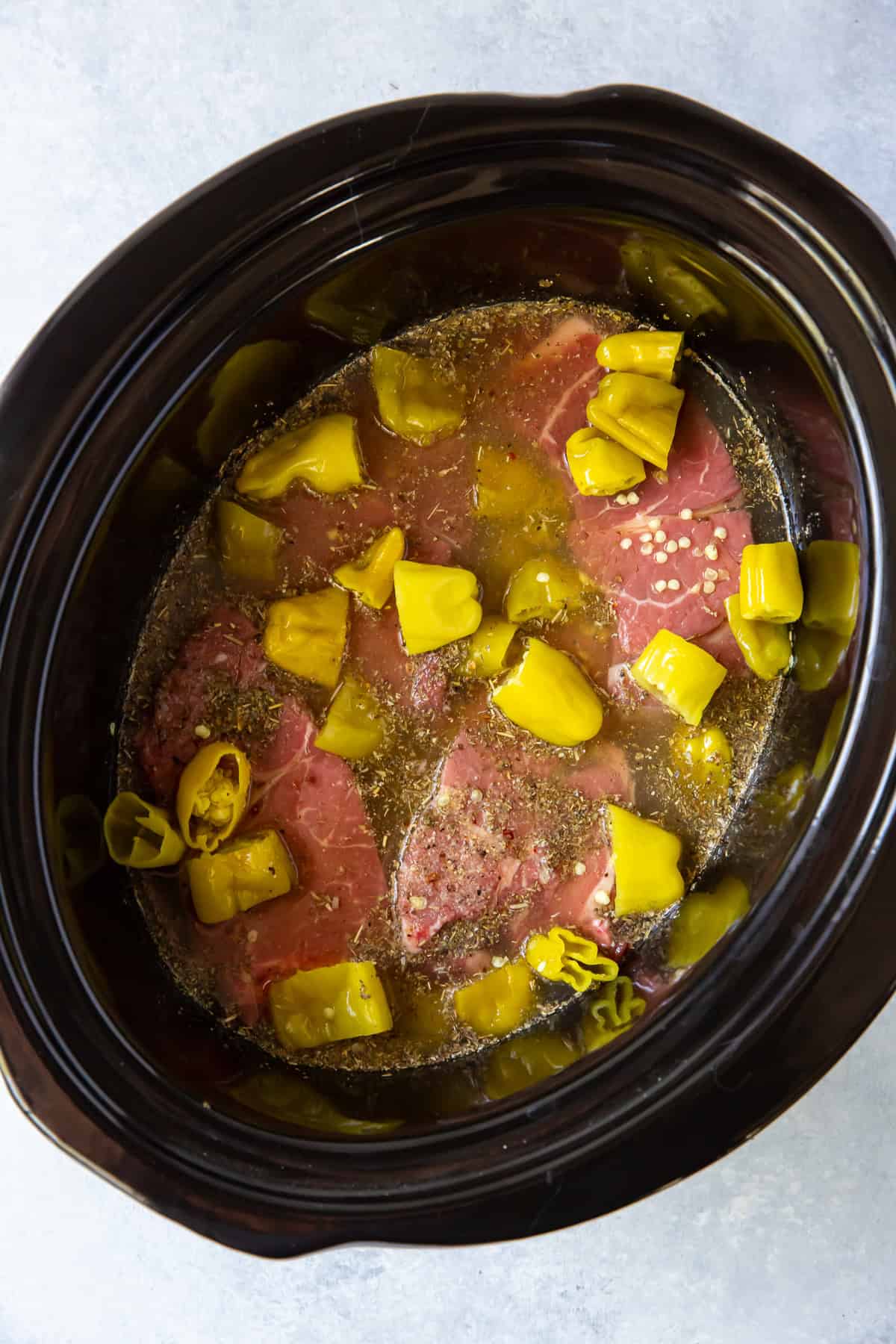 Roughly chopped pepperoncini on top of chunks of chuck roast in broth in a slow cooker insert.