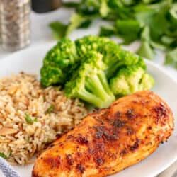 A close up of cooked chicken breast on a plate with rice and broccoli.