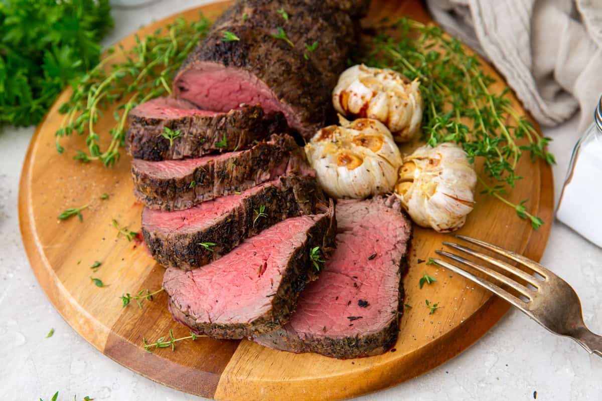 Sliced beef tenderloin and roasted garlic on a wood board.