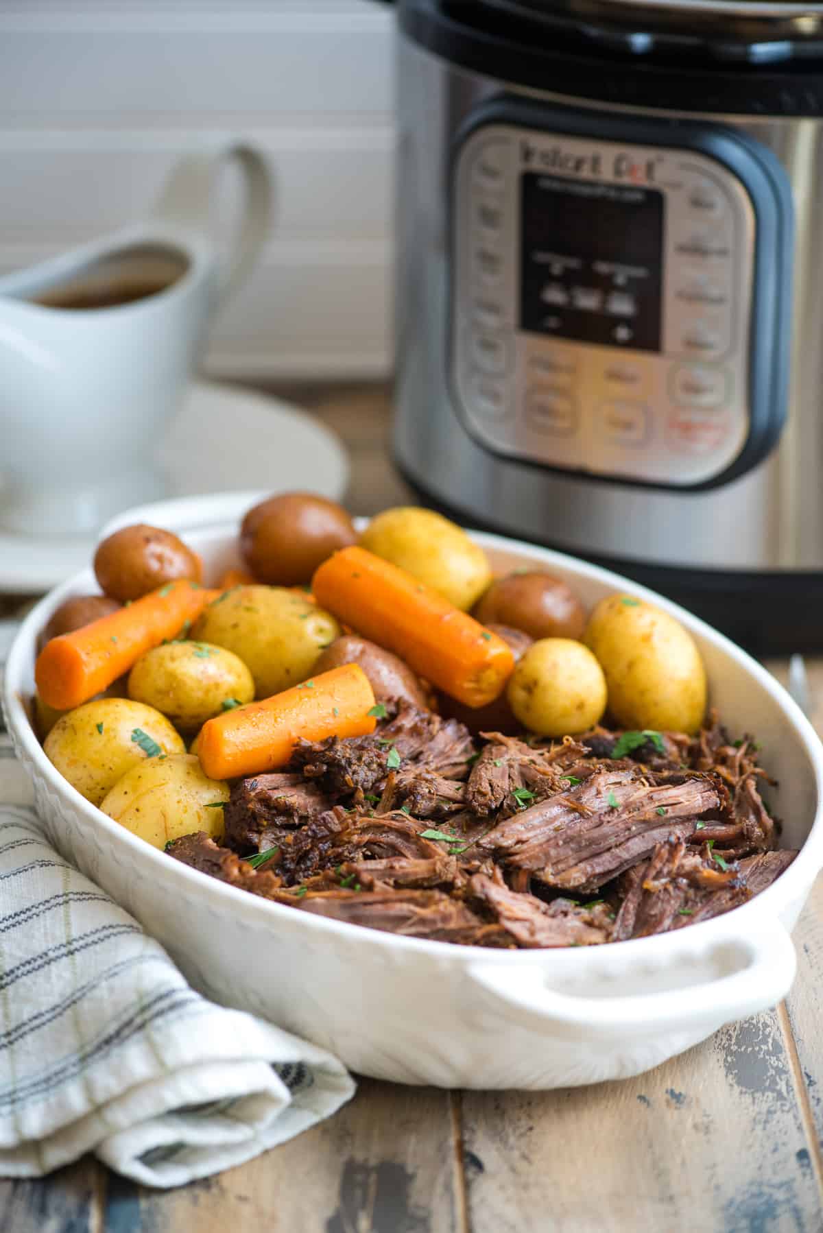 Pot roast with carrots and potatoes in a white serving dish with an Instant Pot in the background.