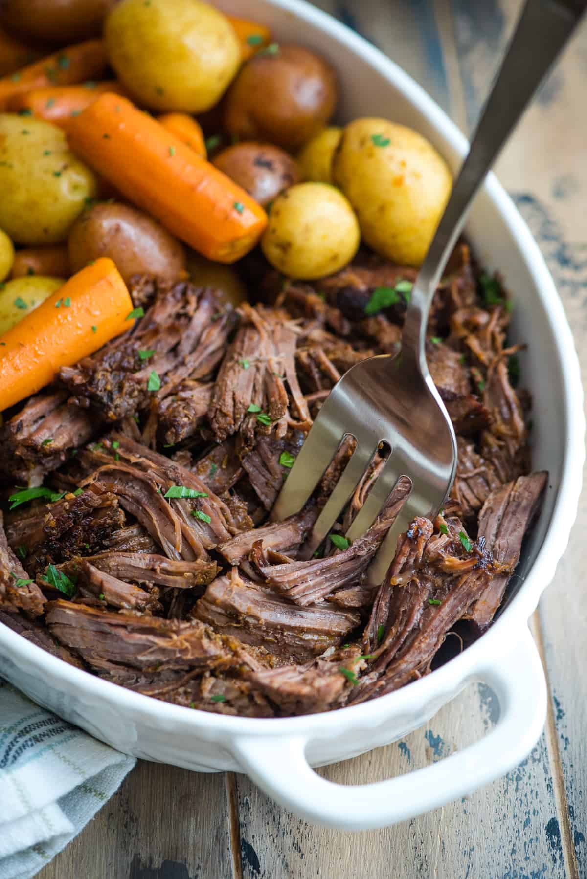 Close up of pot roast with carrots and potatoes in a white serving dish with a fork.