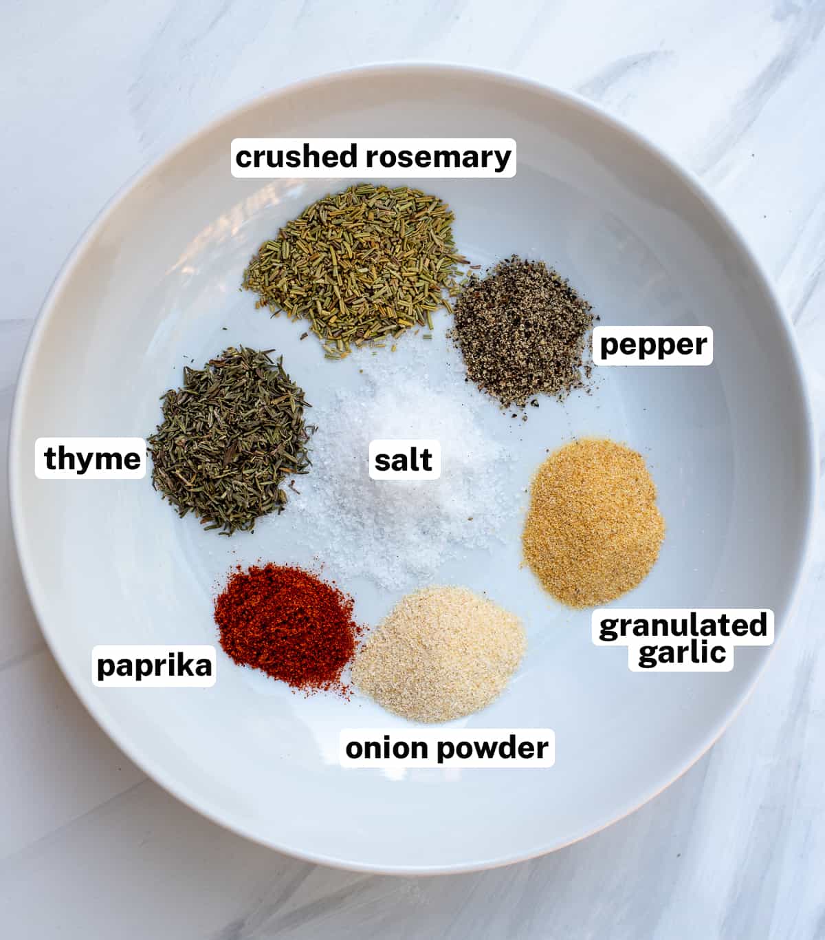 An assortment of dried herbs for Pot Roast Dry Rub on a white dish.