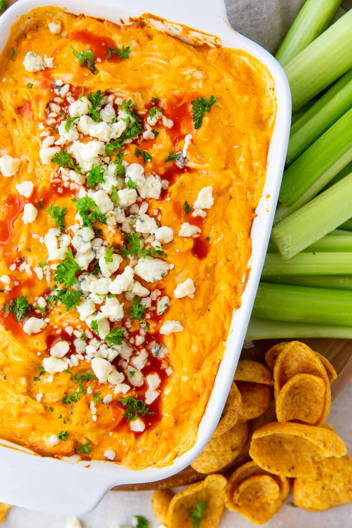 Buffalo Chicken Dip garnished with blue cheese and Frank's Red Hot in a baking dish next to celery and Frito's Scoops.