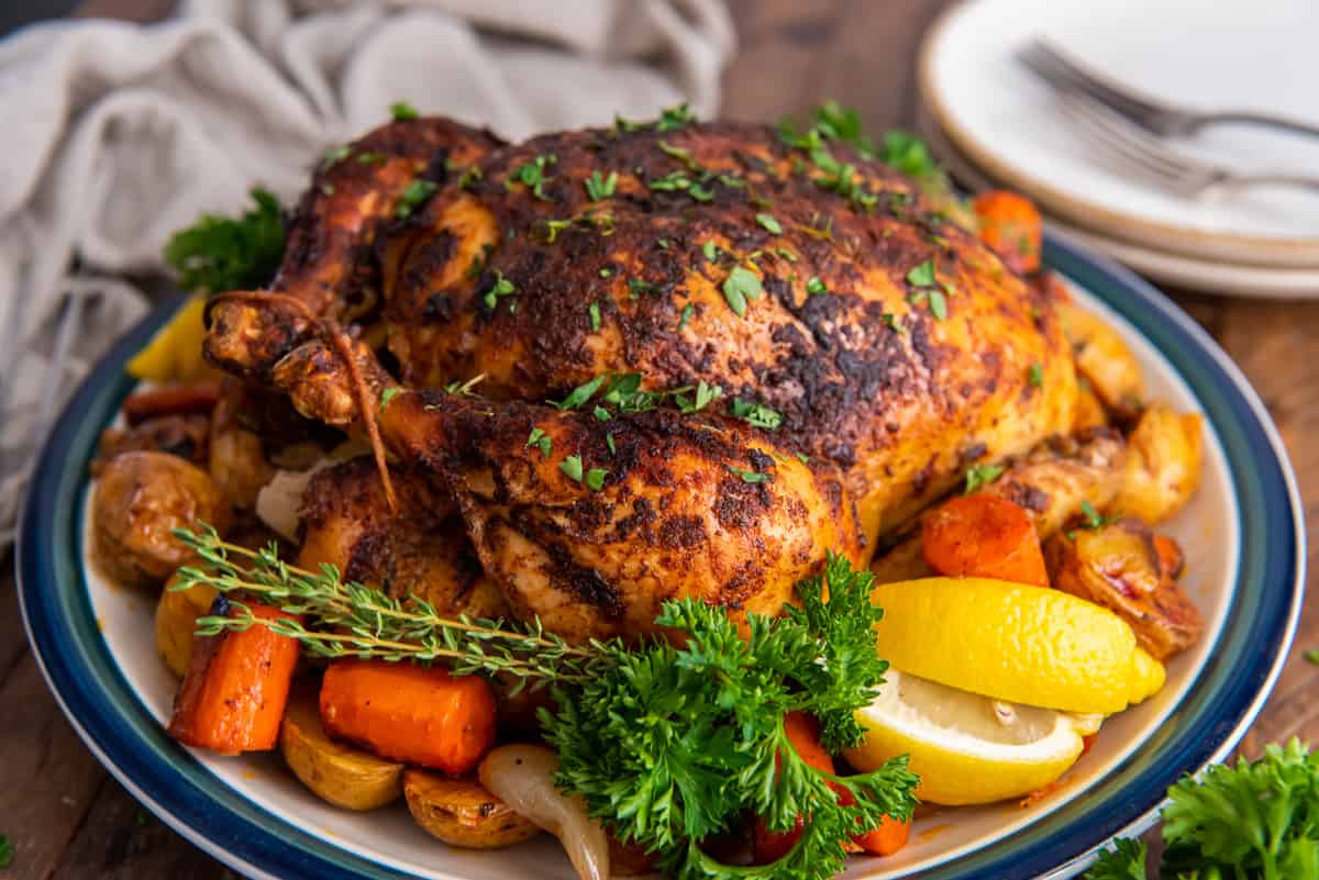 A whole roasted chicken on a platter with vegetables, fresh herbs, and lemon wedges.