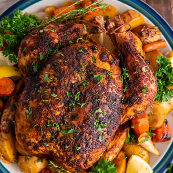 An over the top shot of a roasted whole chicken on a platter with vegetables, fresh herbs, and lemon wedges.