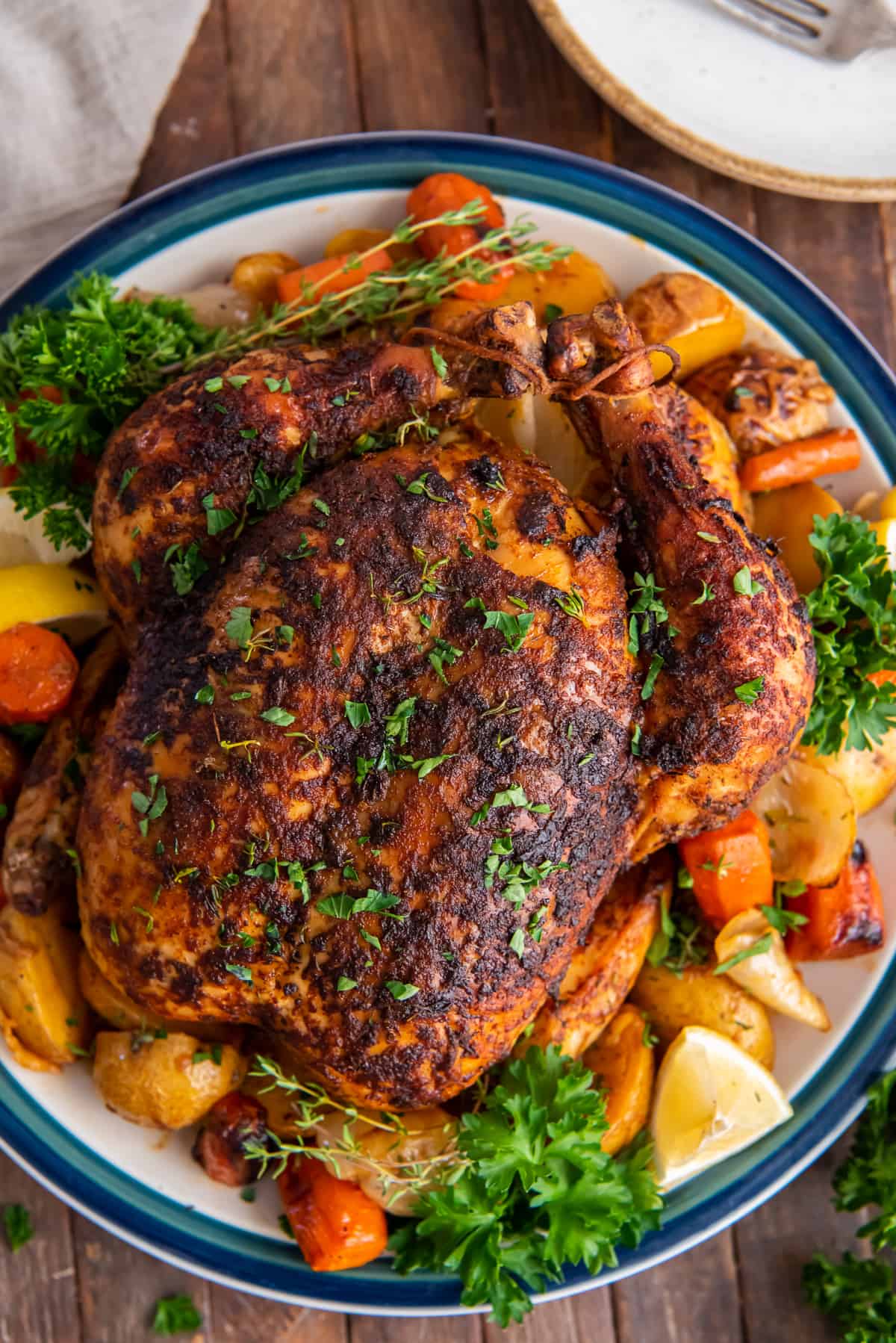 An over the top shot of a roasted whole chicken on a platter with vegetables, fresh herbs, and lemon wedges.