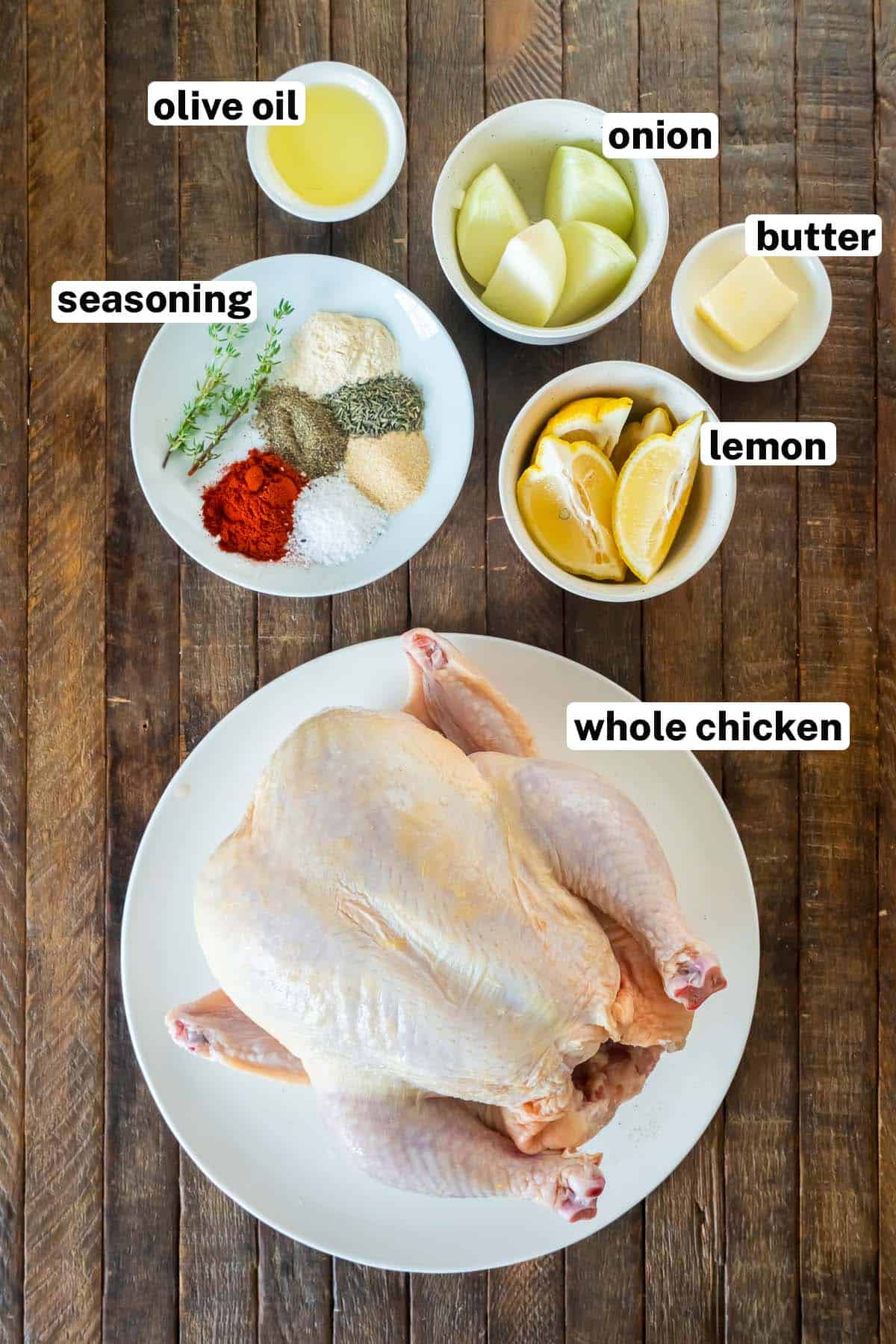 A whole chicken, seasoning, lemon wedges, onion, and oil on a wood surface with text.