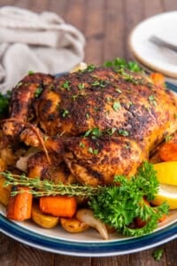 A crispy roast chicken on a platter with carrots and potatoes.