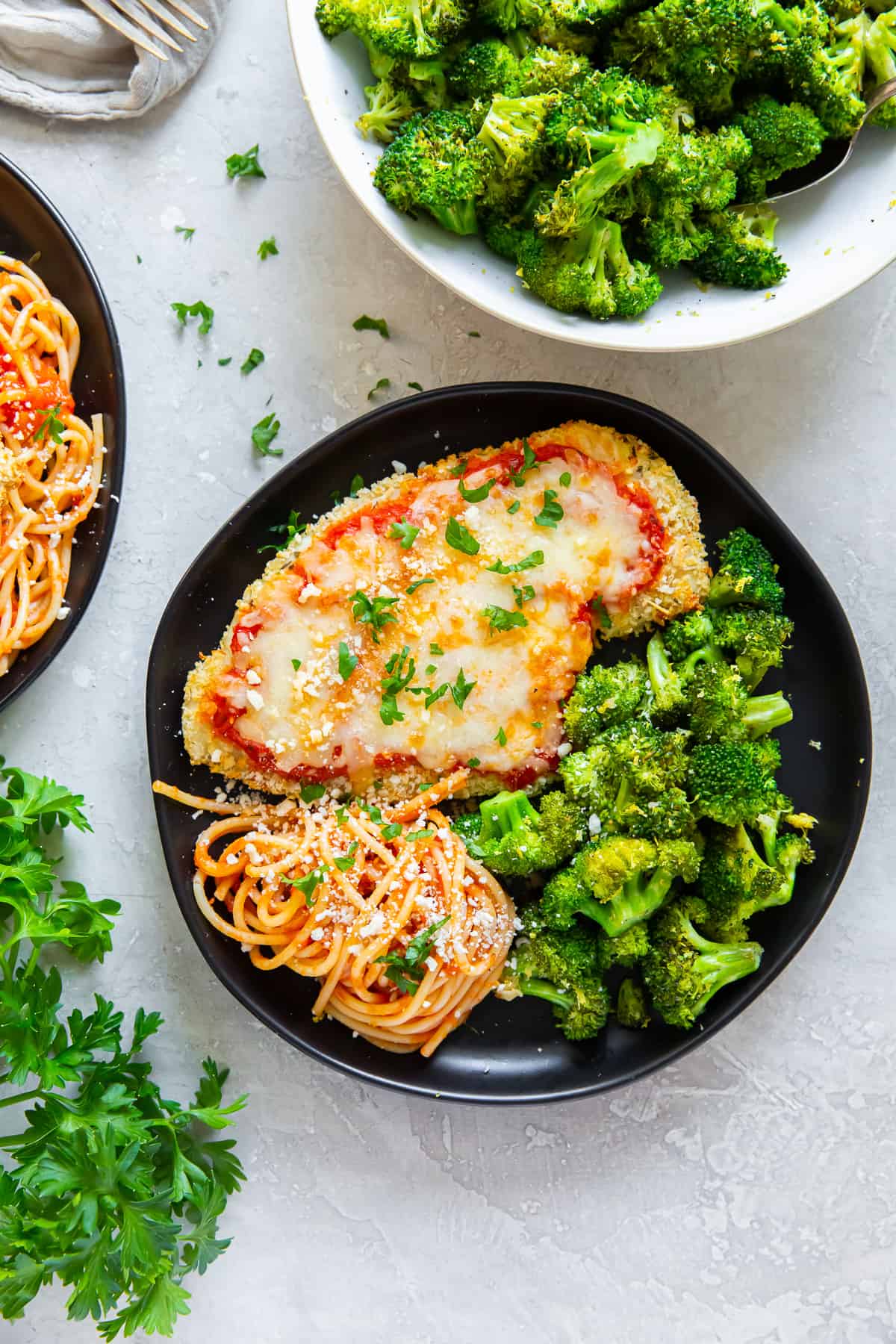 Chicken parmesan on a black plate with pasta and broccoli.