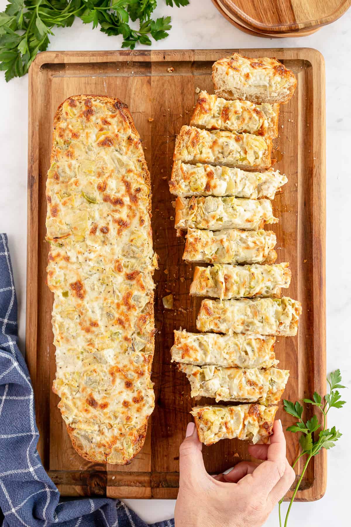 A hand reaching for a slice of Cheesy Artichoke Bread on a cutting board.