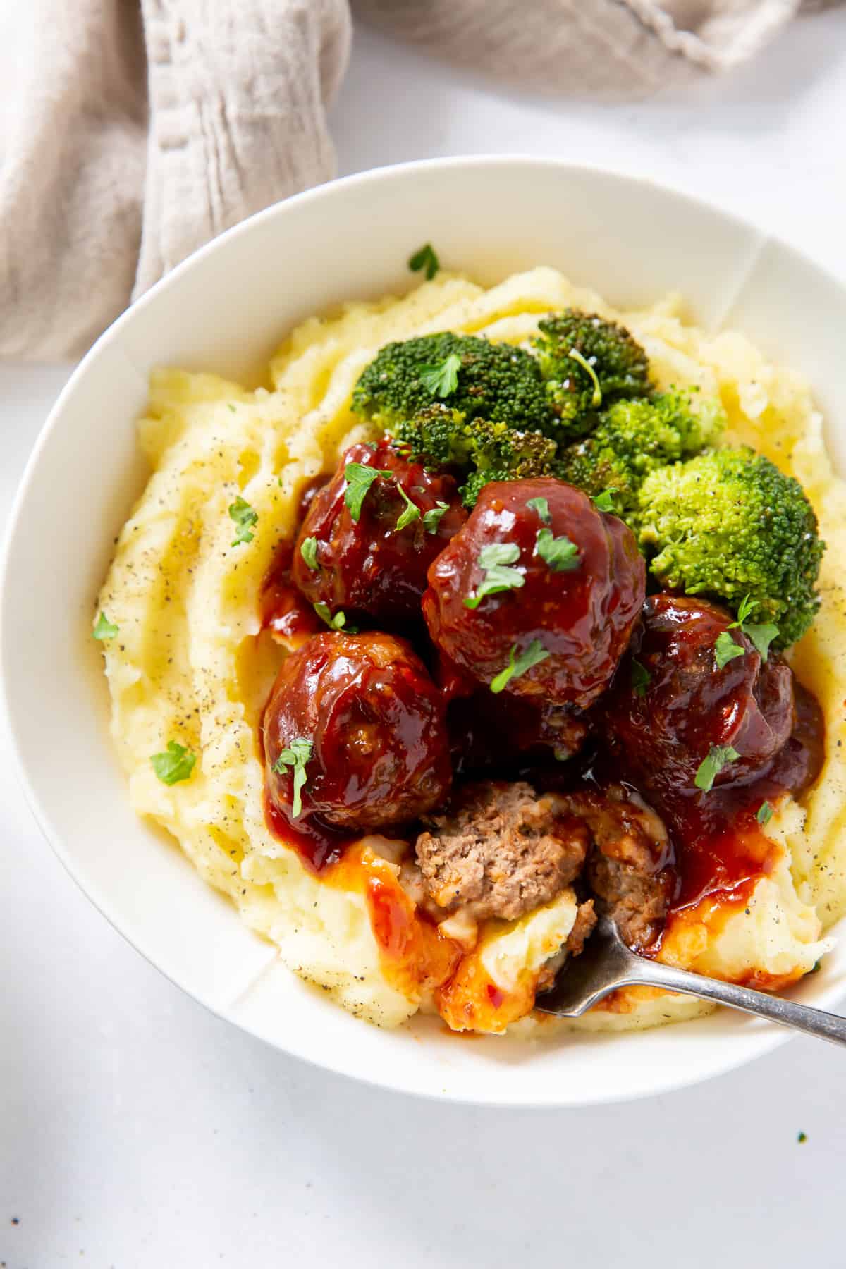A fork breaking into a meatball in a bowl with mashed potatoes.