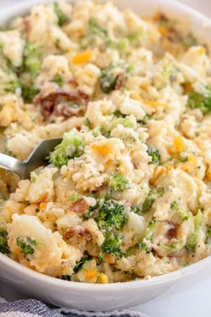 A spoon scoops mashed potatoes with broccoli from a white bowl.