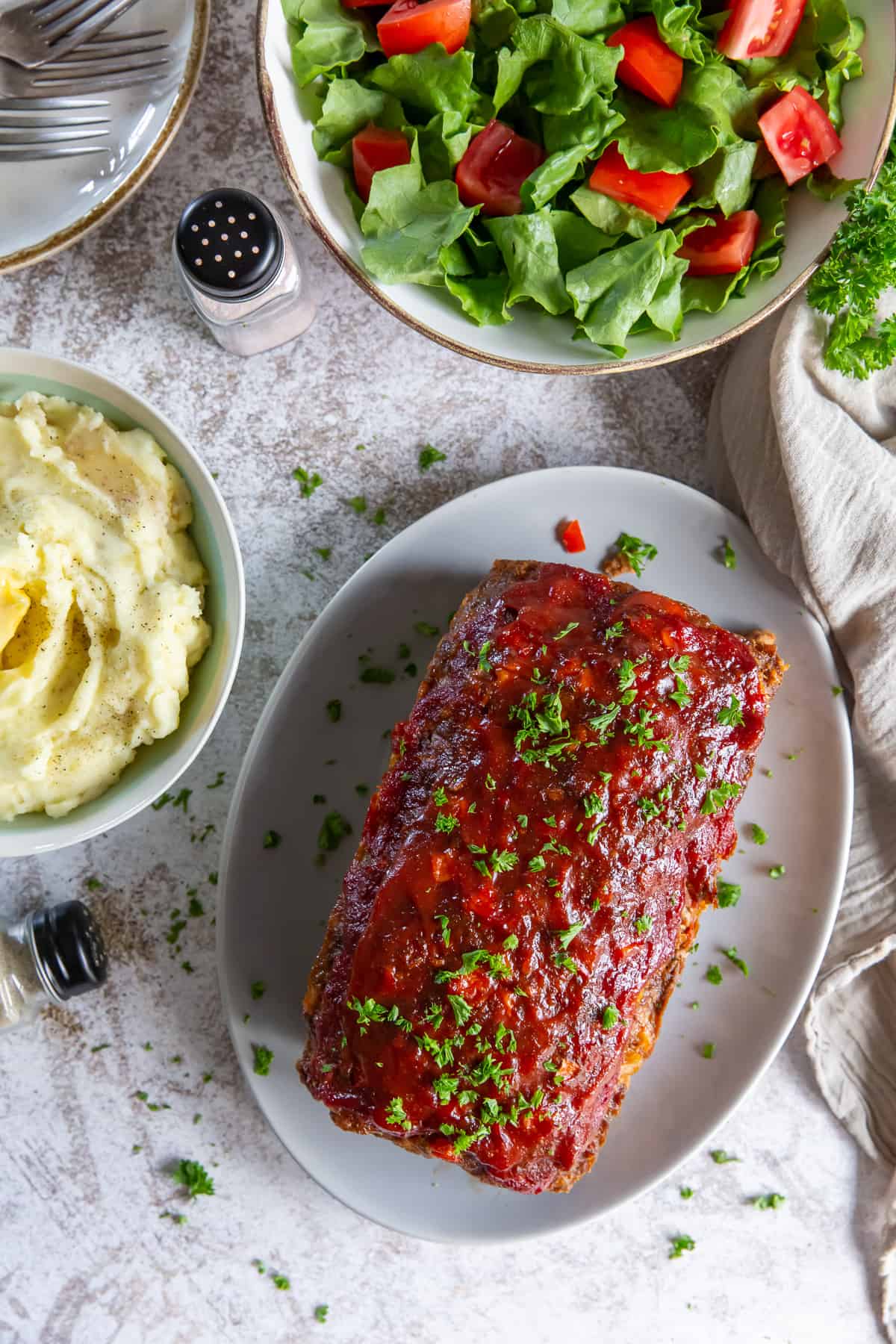 A glazed meatloaf on a platter next to a salad and mashed potatoes.