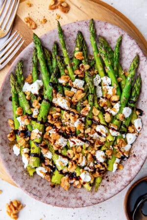 A top down shot of asparagus with balsamic glaze, goat cheese, and walnuts on a platter.