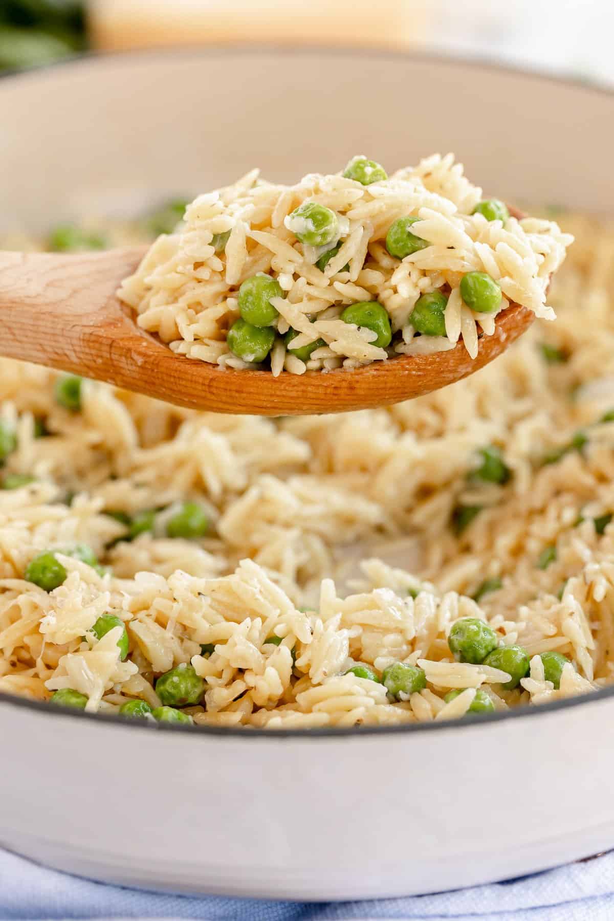 A wooden spoon lifting a scoop of orzo with peas and parmesan from a skillet.