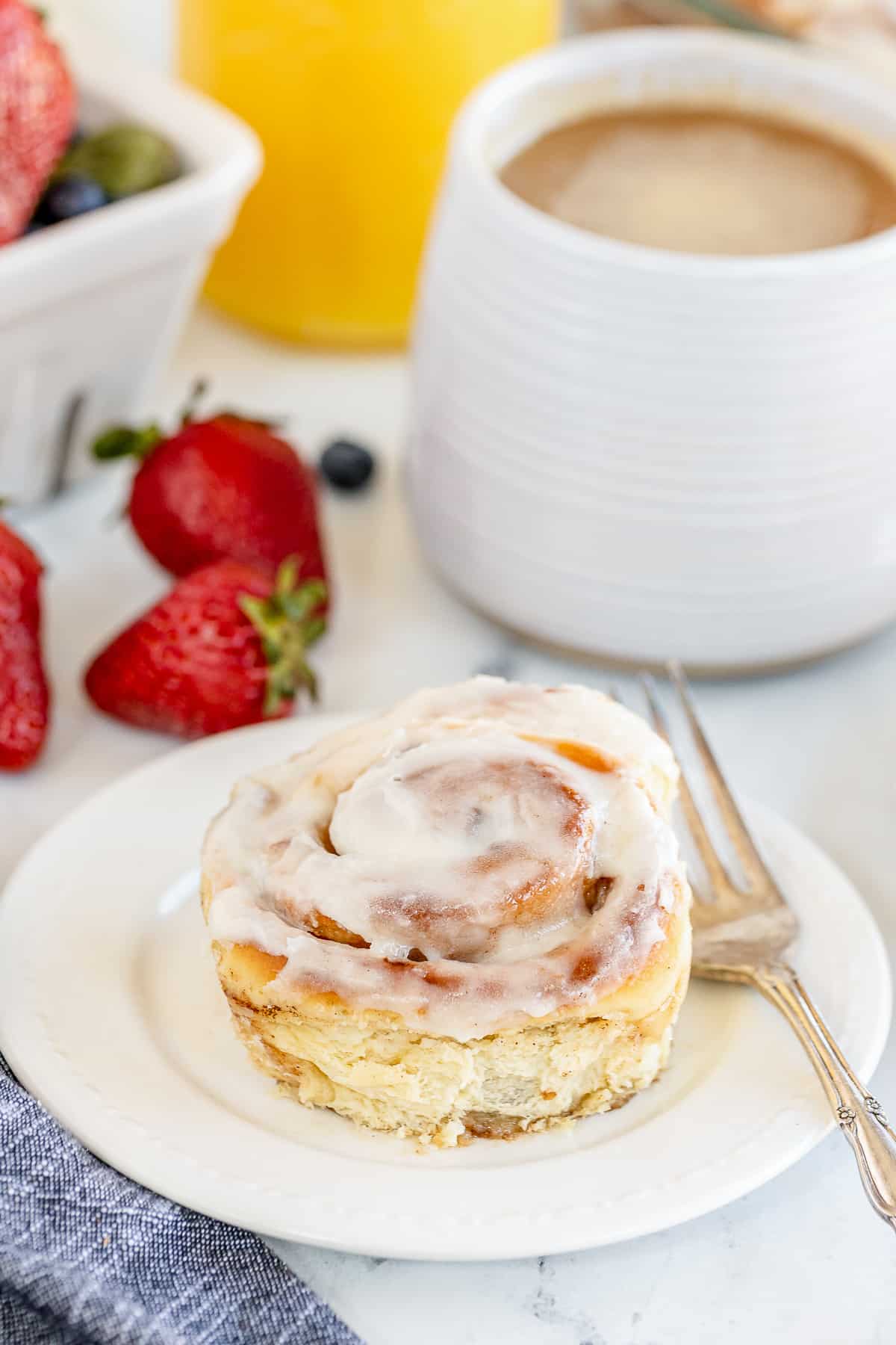 A cinnamon roll on a plate next to a cup of coffee and strawberries.