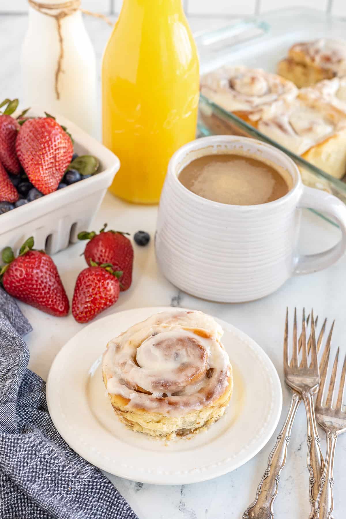 A cinnamon roll on a plate with a cup of coffee and orange juice in the background.