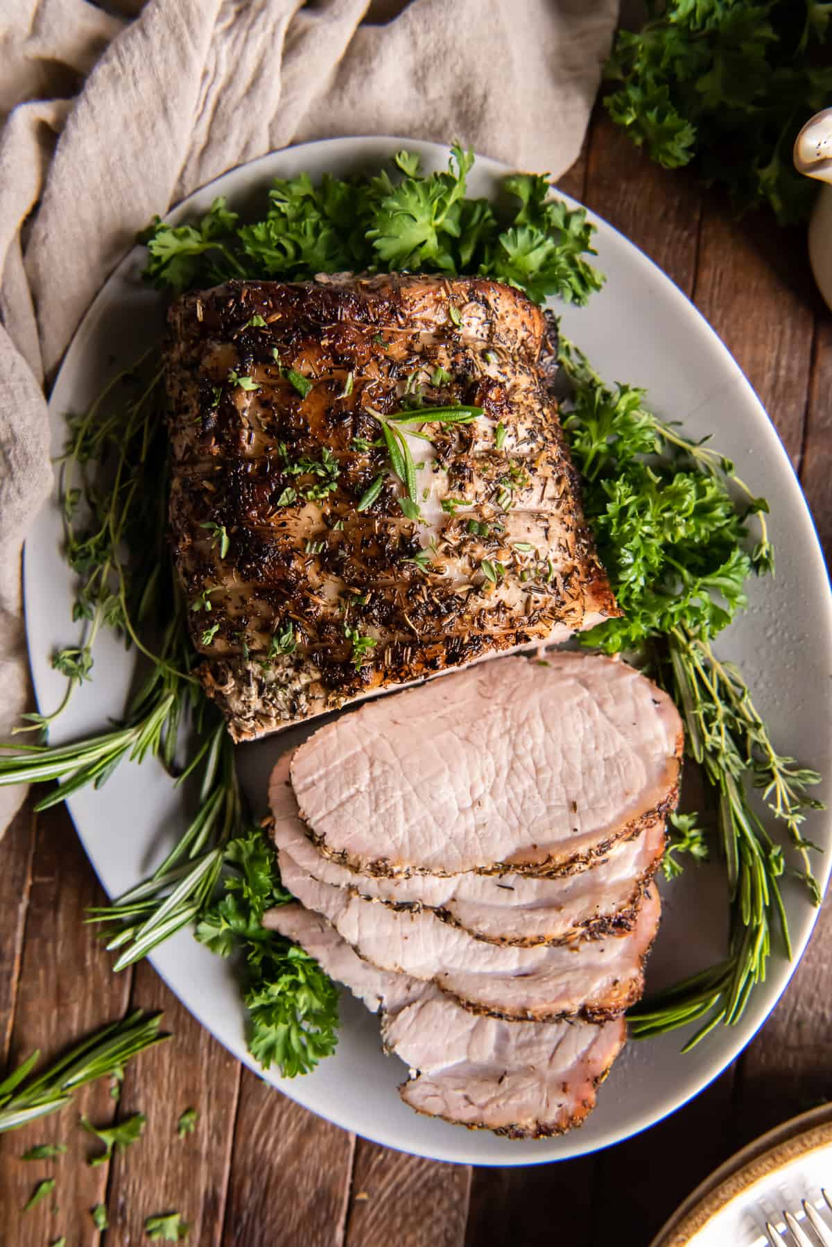 A top down shot of a sliced pork loin roast on a white platter with herbs.