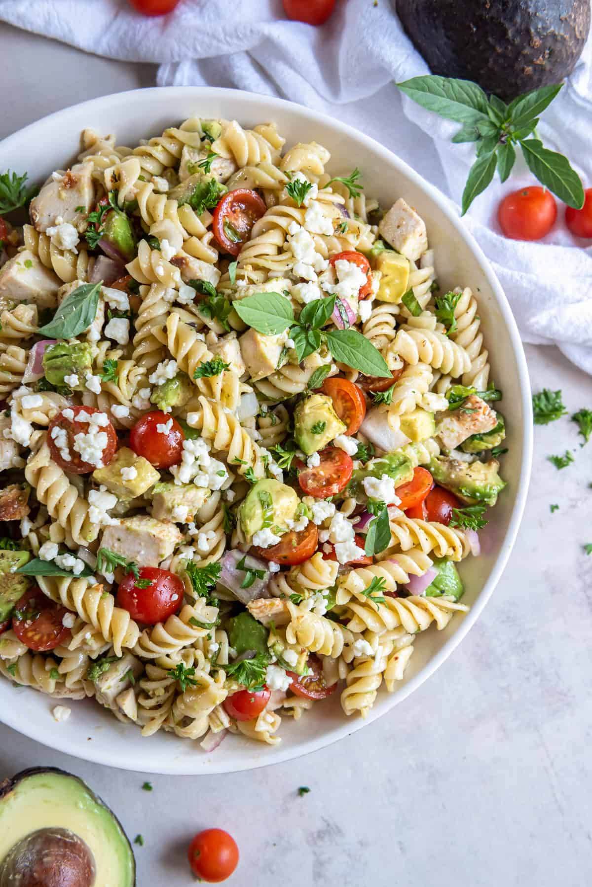Pasta salad with avocado, chicken, and tomatoes in a white bowl with spoons.
