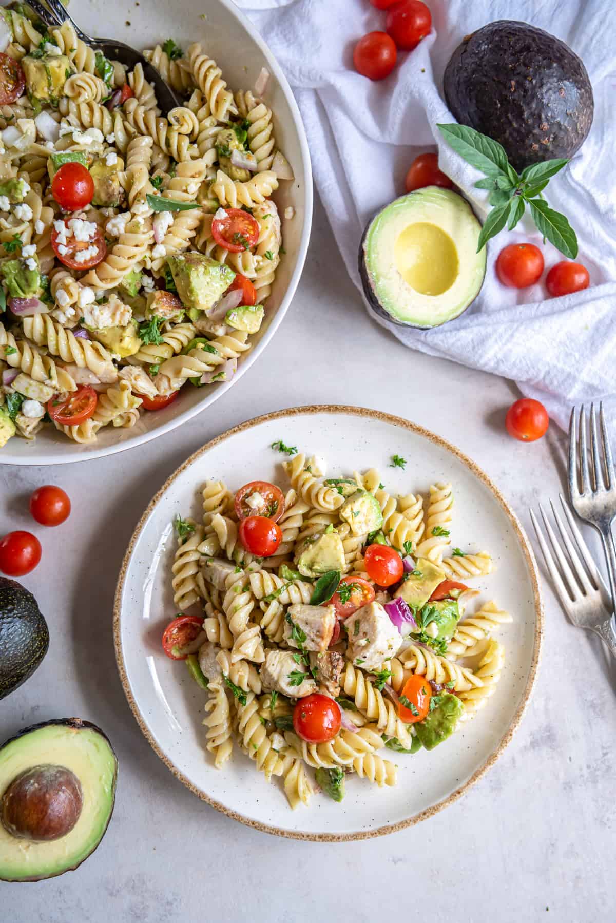 Chicken pasta salad in a serving bowl and on a plate next to avocados and cherry tomatoes.