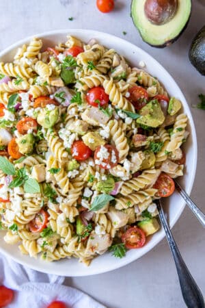 A white bowl filled with chicken avocado pasta salad and salad spoons.