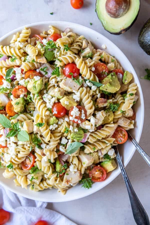 A white bowl filled with chicken avocado pasta salad and salad spoons.