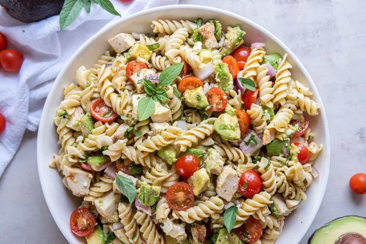 A top down shot of chicken avocado pasta salad with cherry tomatoes and basil in a white bowl.