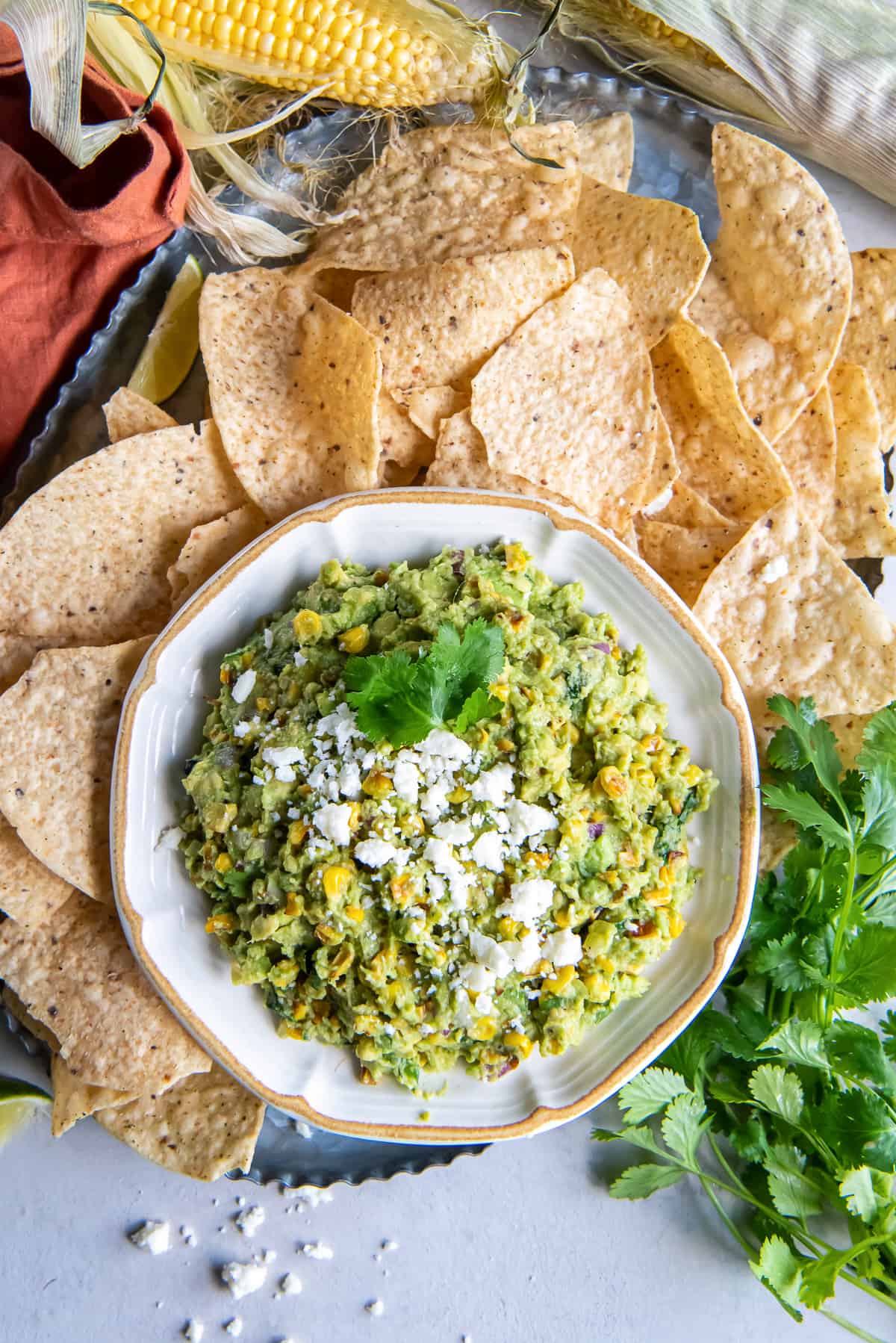 A top down shot of a bowl of corn guacamole surrounded by tortilla chips and sweet corn.