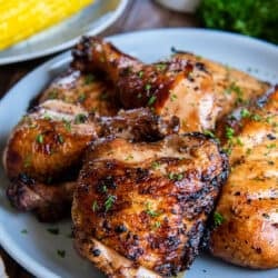 Grilled chicken on a white plate with corn on the cob in the background.