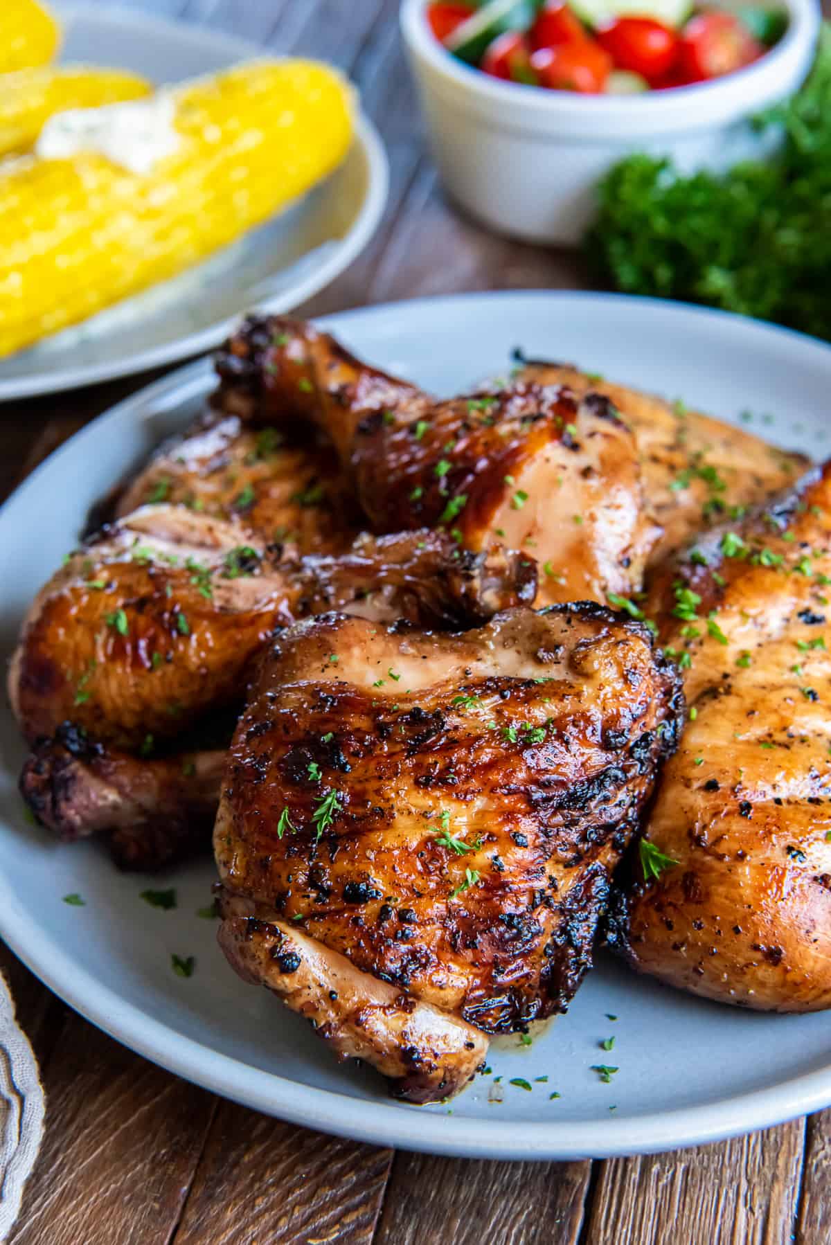 Grilled chicken on a white plate with corn on the cob in the background.