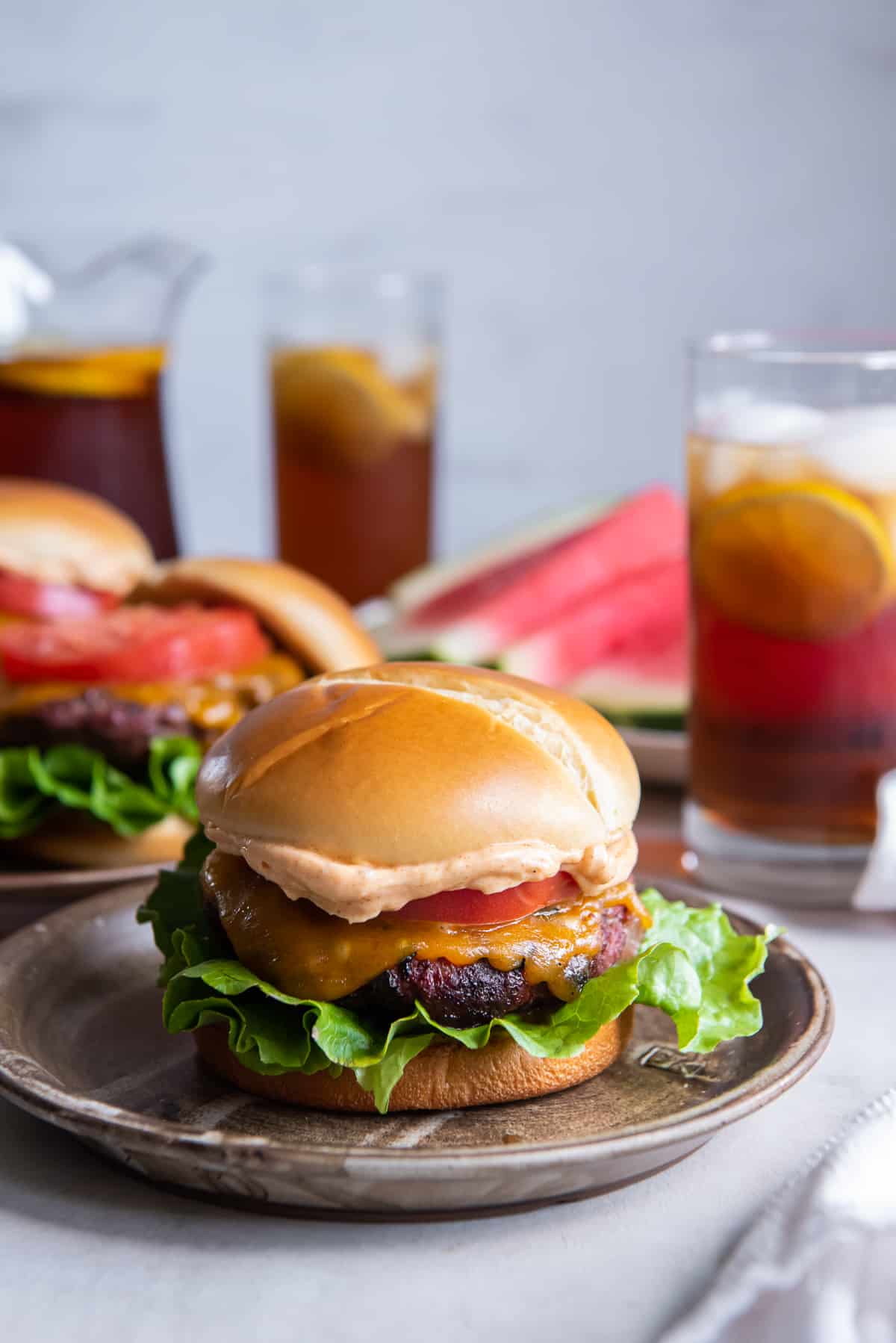 A Ranch Burger on a wood plate in front of iced tea and watermelon.
