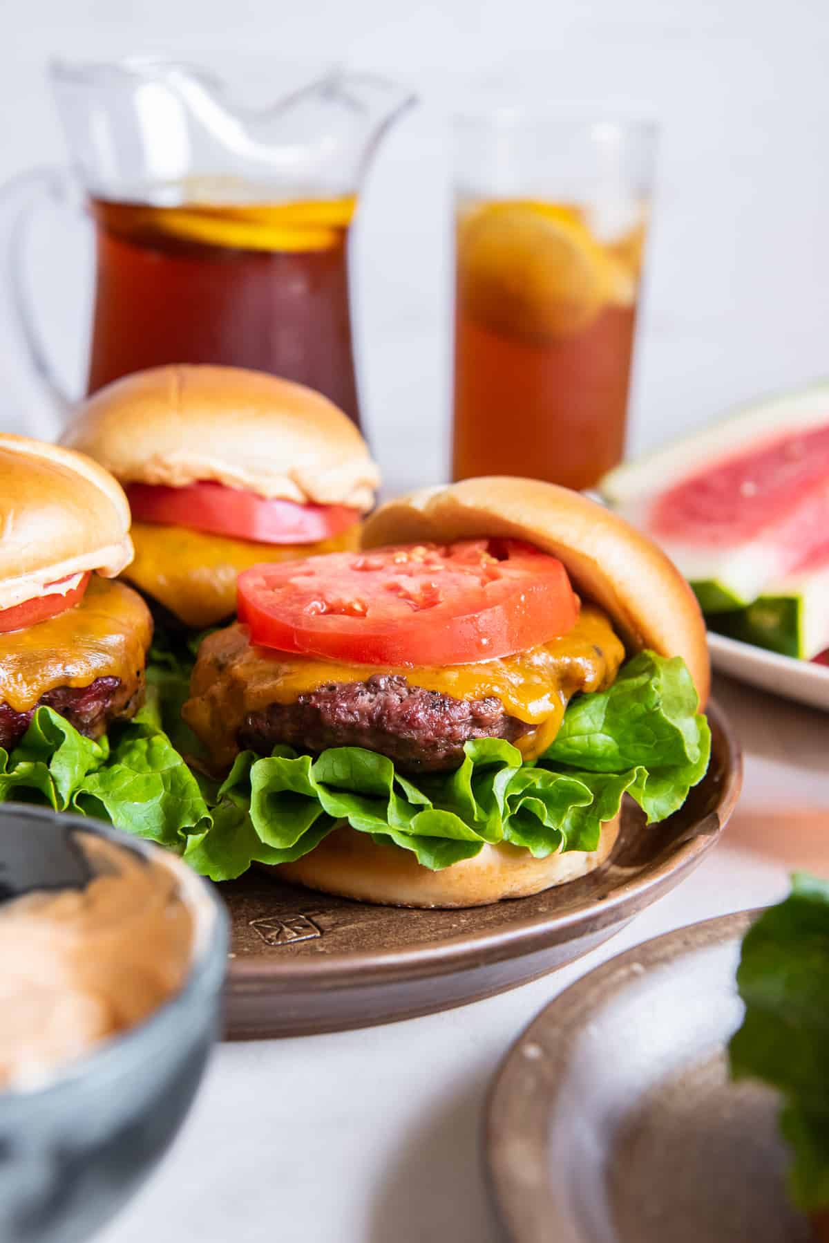 A burger  with lettuce and tomato in front of a pitcher of iced tea.