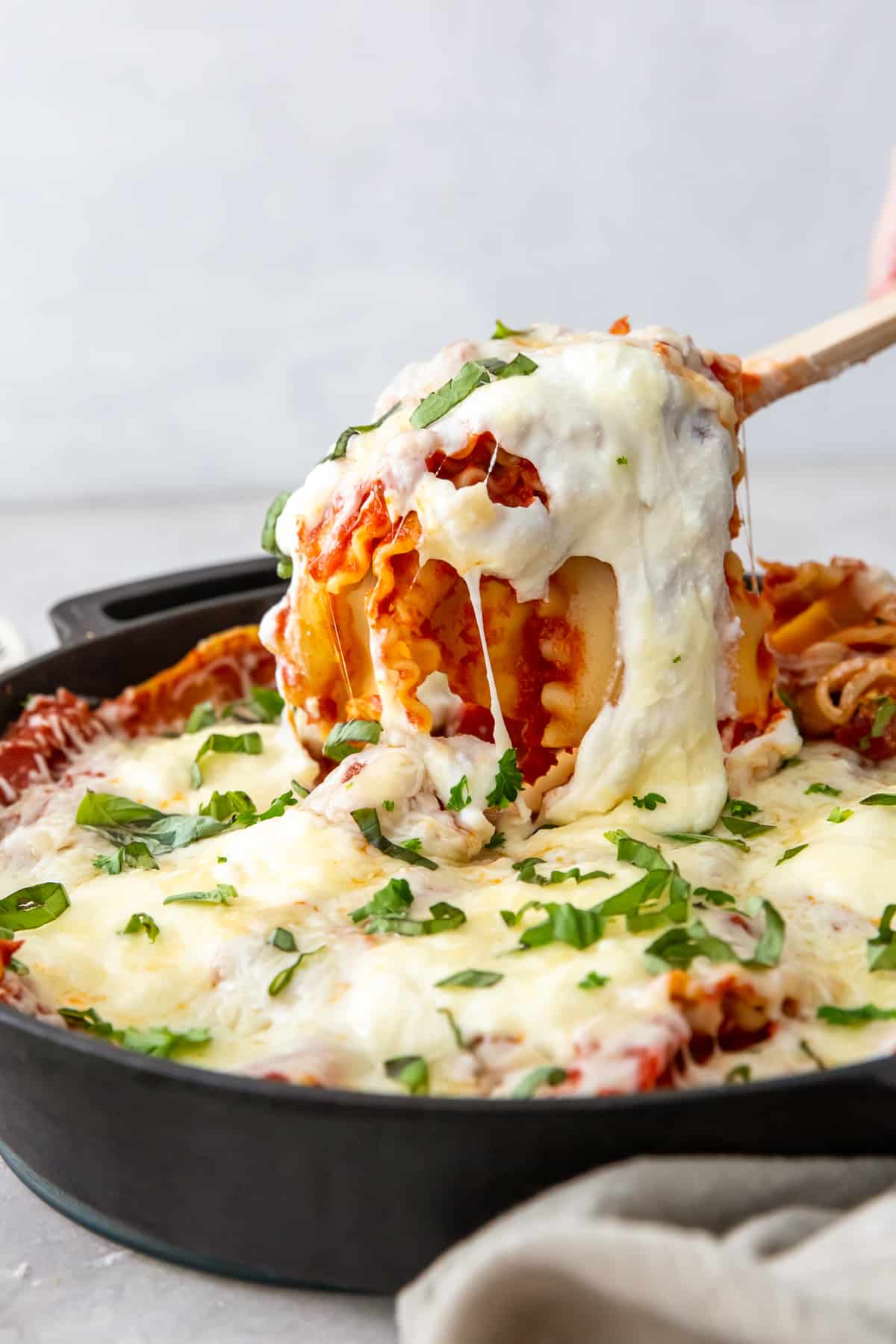 A wooden spoon scoops lasagna from a skillet.