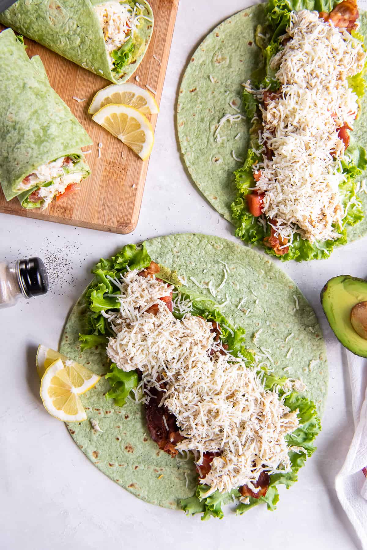 Two spinach wraps laying flat and topped with avocado, chicken and cheese. Lemon wedges and halved avocados lie nearby.