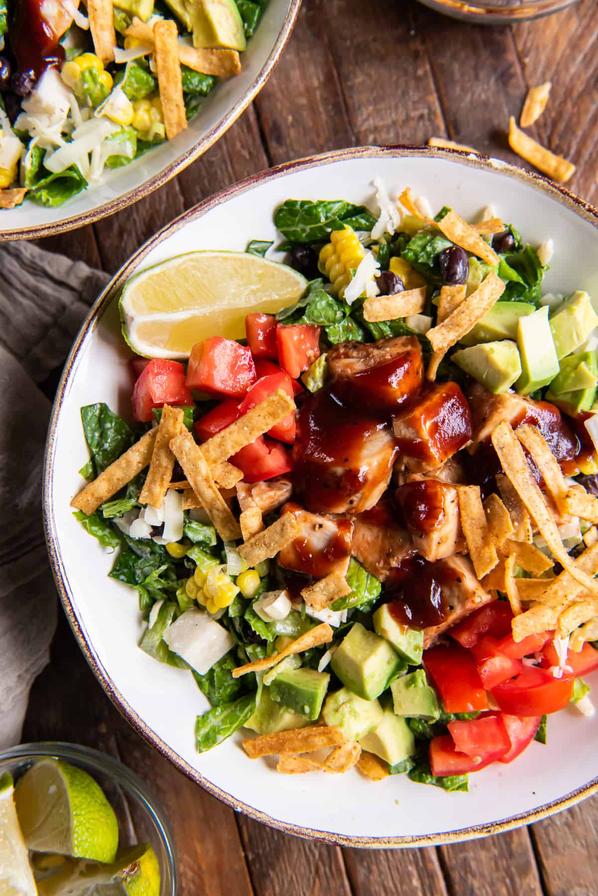 A top down shot of a salads with chicken, bbq sauce, tomatoes on a dark wood board with lime wedges next to it.