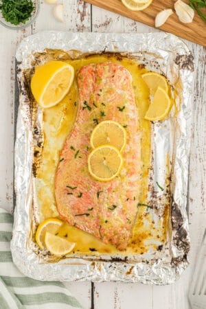A top down shot of a large baked salmon fillet topped with butter and garlic sauce and lemon slices on a large sheet of foil.