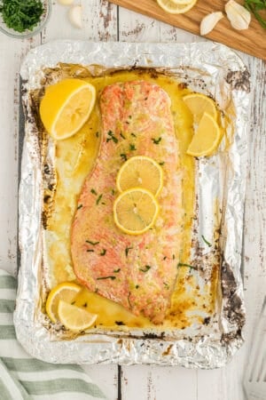 A top down shot of a large baked salmon fillet topped with butter and garlic sauce and lemon slices on a large sheet of foil.