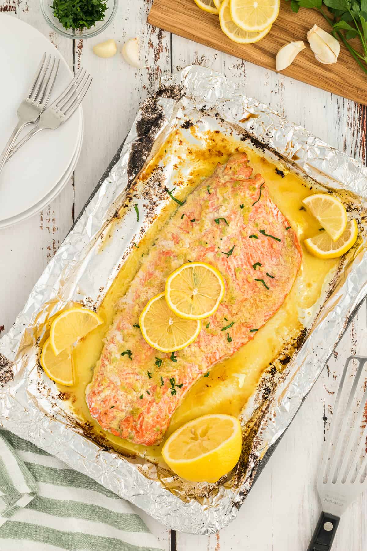 A large salmon fillet with garlic butter sauce a lemon slices on a foil lined baking sheet next to a striped cloth and a cutting board.