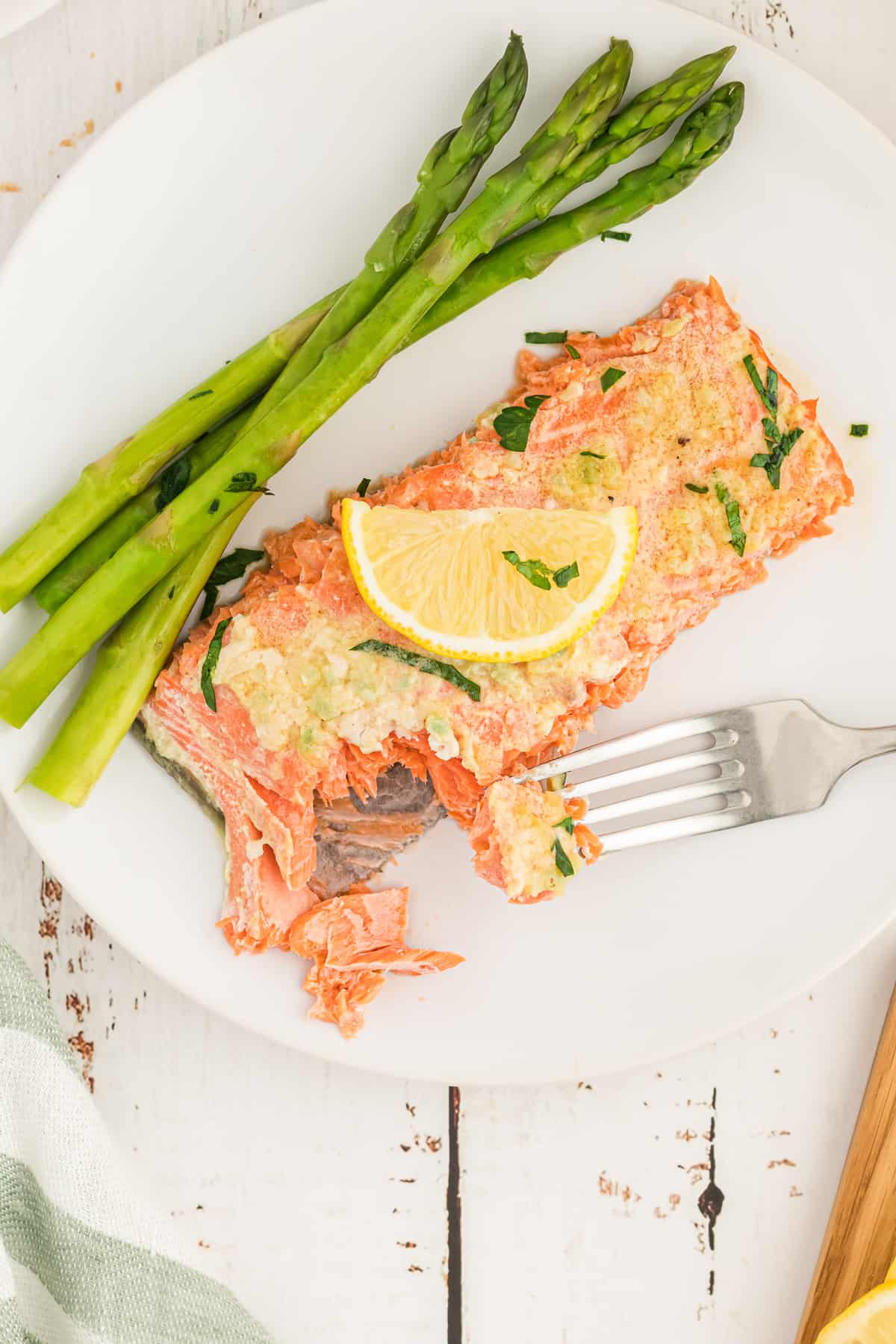 A fork breaking into a slice of salmon topped with a slice of lemon on a white plate with asparagus.