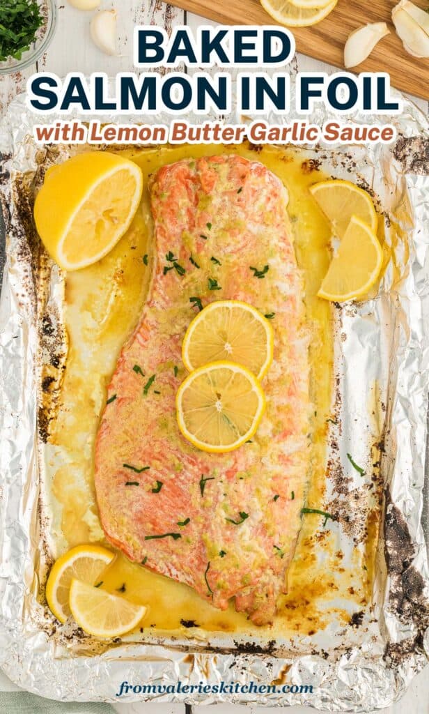 A top down shot of a large baked salmon fillet topped with butter and garlic sauce and lemon slices on a large sheet of foil with text.