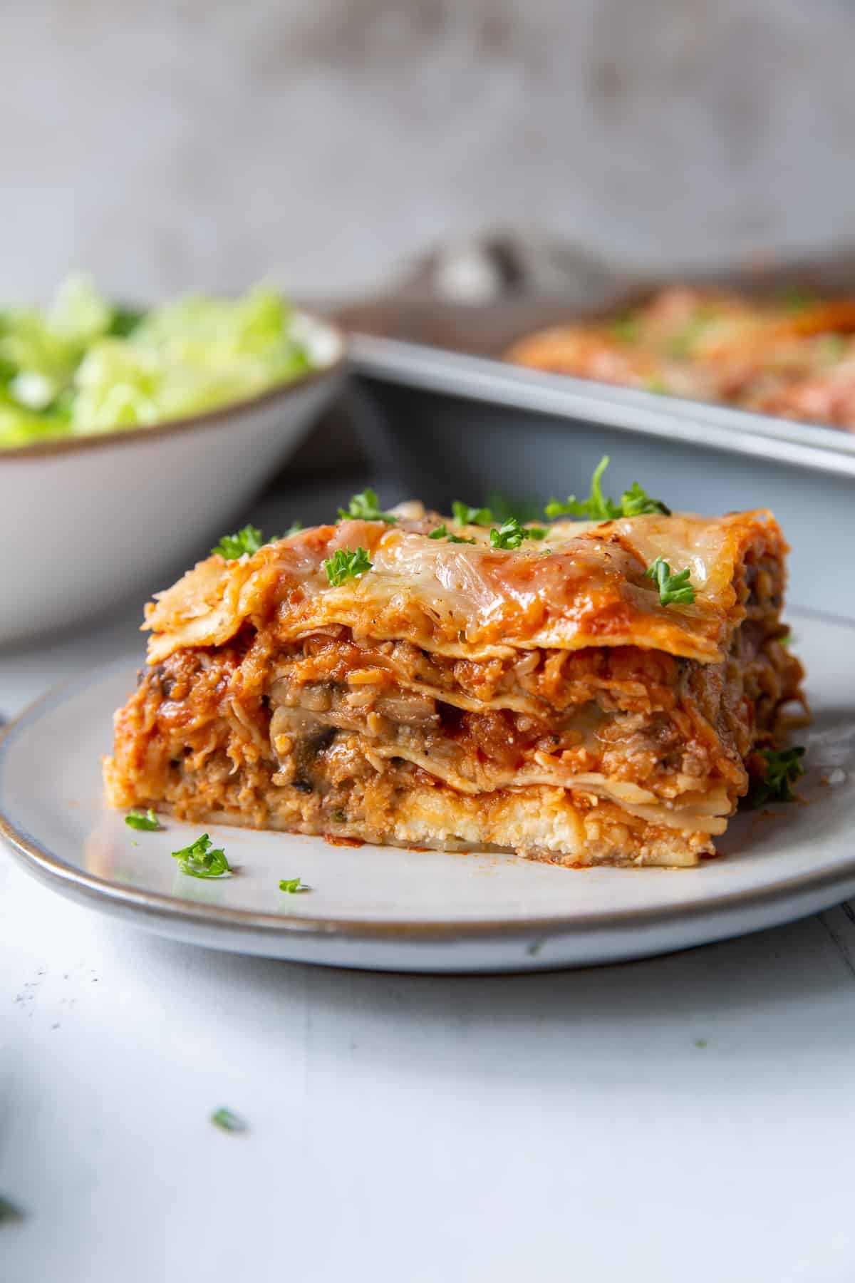 A close up of a slice of lasagna on a small white plate with a salad bowl and baking dish in the background.