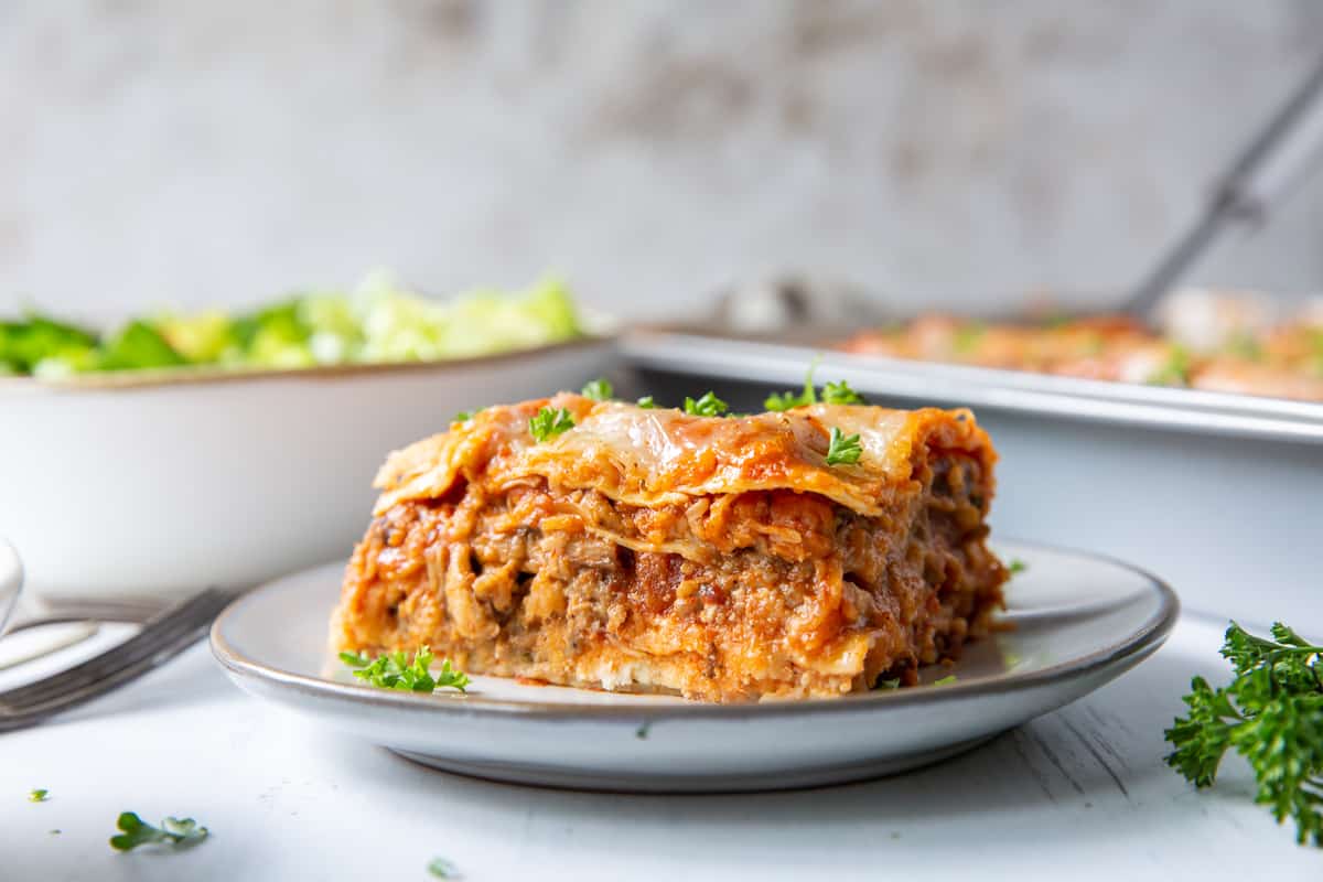 A slice of lasagna on a white plate with a salad and baking dish in the background.