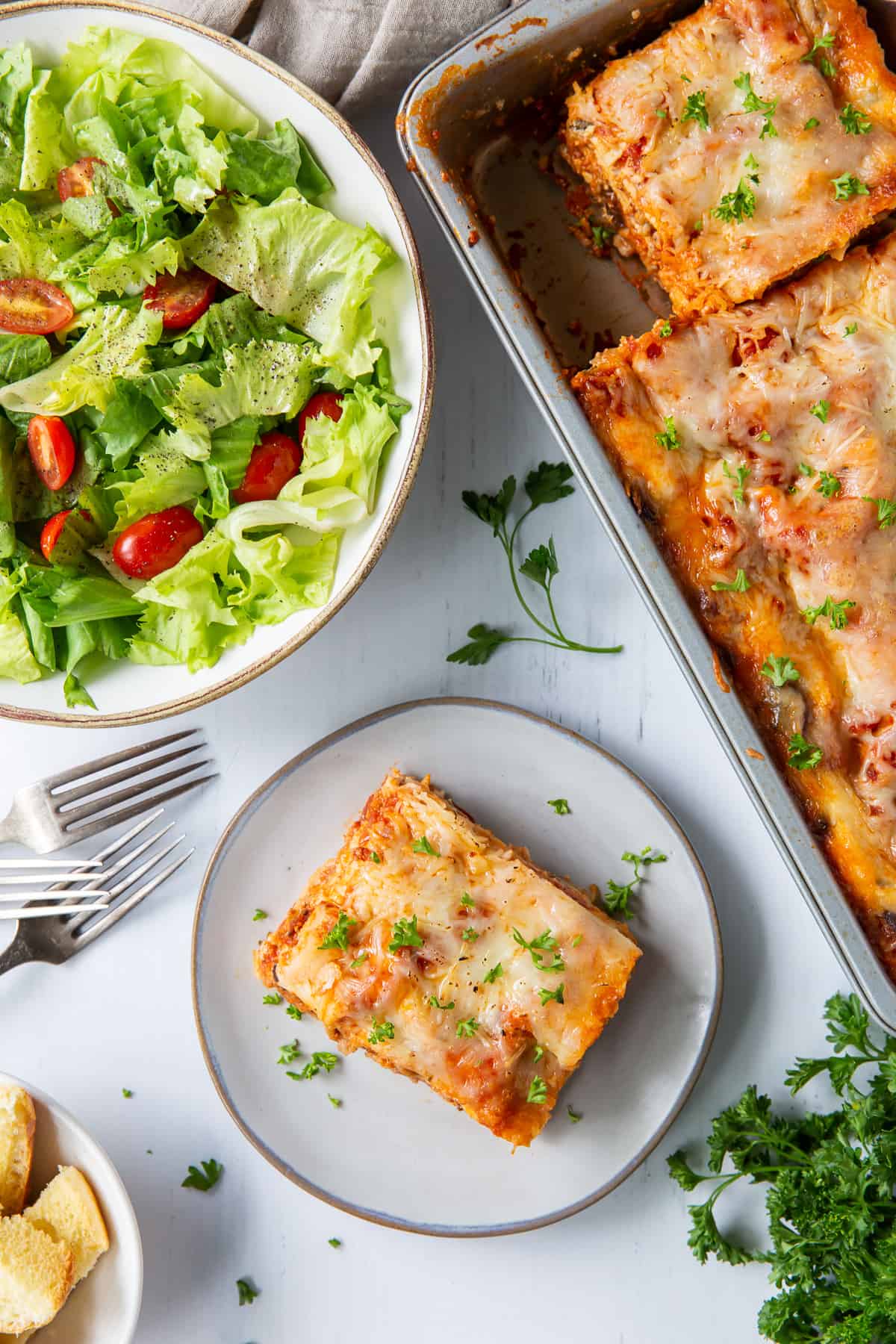 A top down shot of a lasagna in a metal pan next to a green salad and a slice of lasagna on a small white plate.