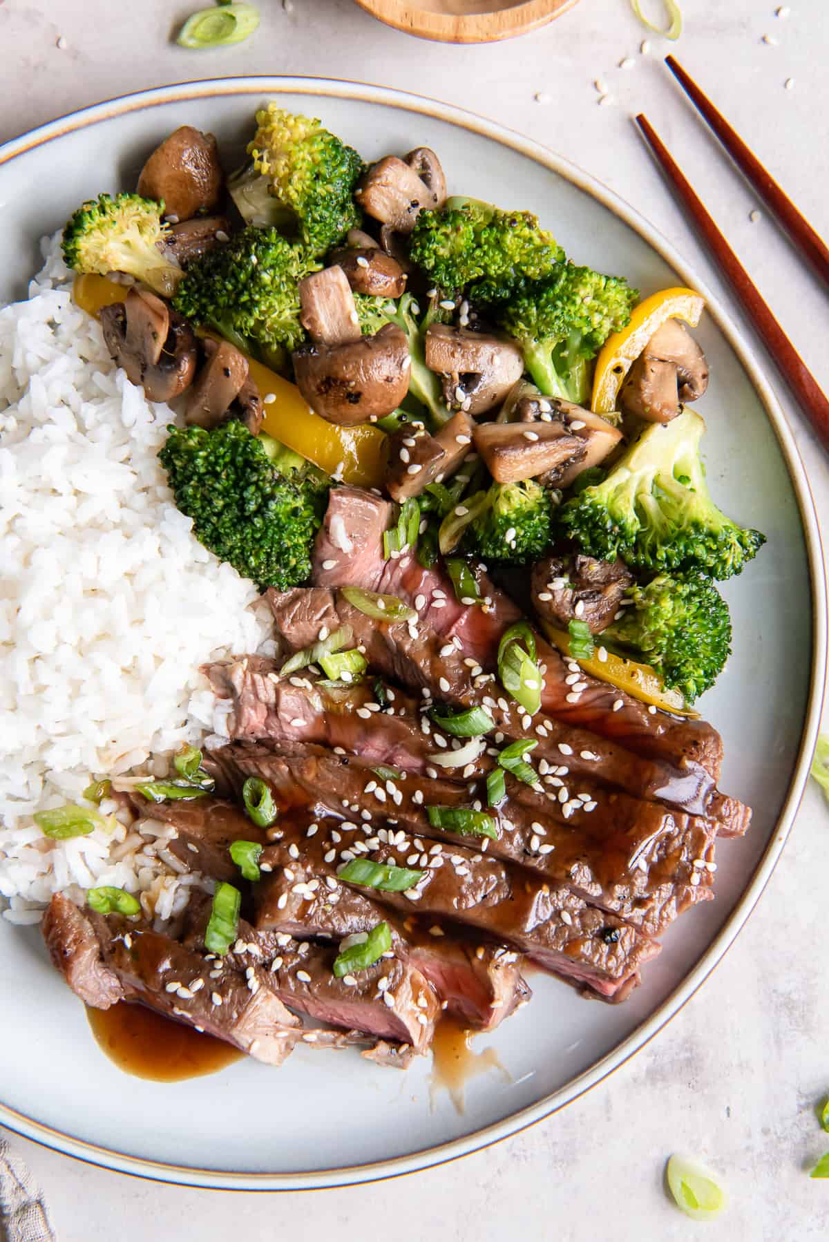 Slices of grilled teriyaki steak on a white plate with rice and stir fried vegetables.