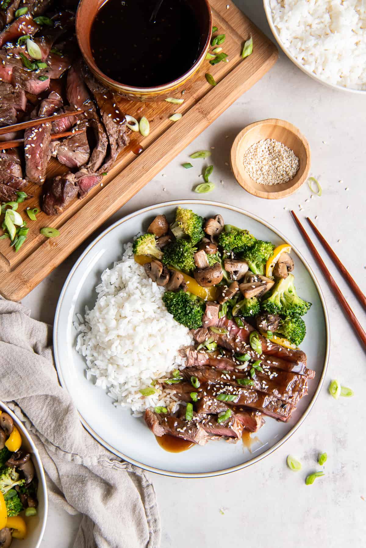Slices of grilled teriyaki steak on a white plate with rice and stir fried vegetables next to a cutting board filled with sliced steak.