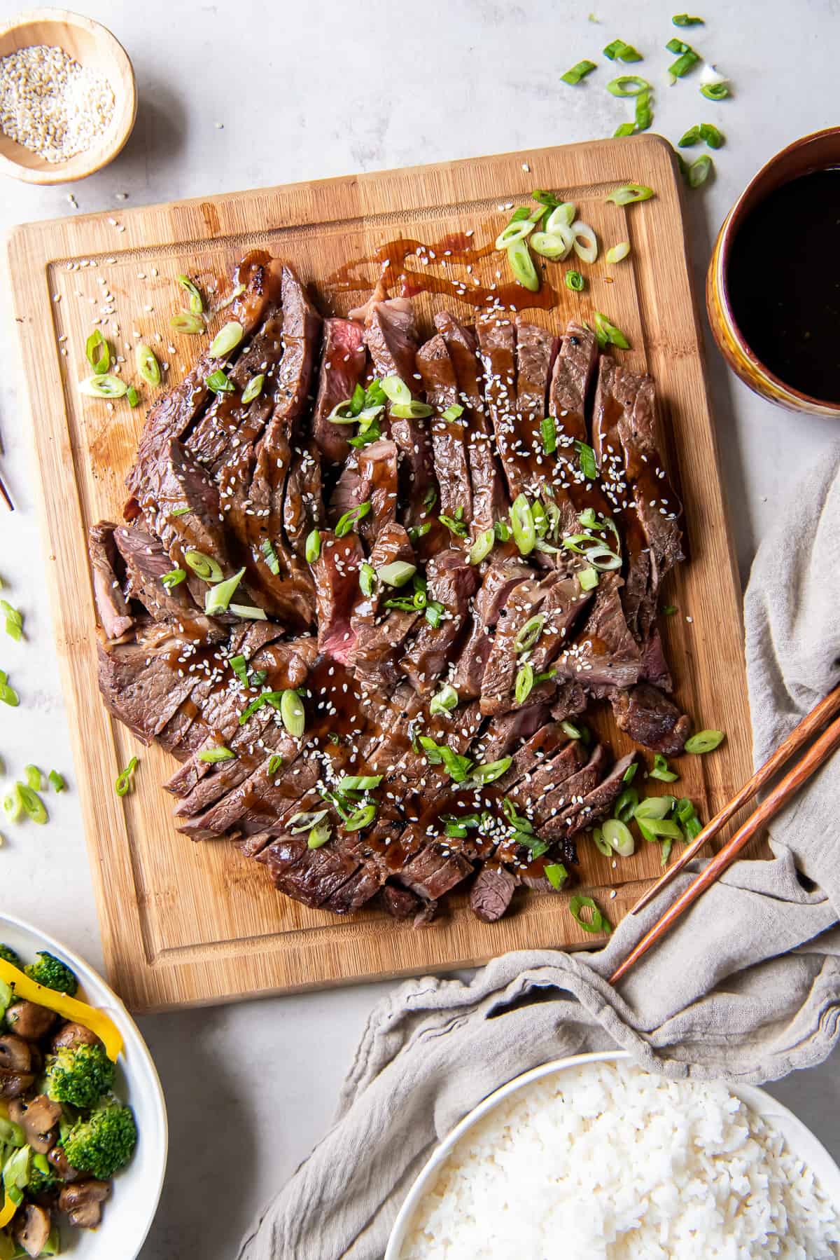 Sliced steak with teriyaki sauce topped with green onions on a wood cutting board.