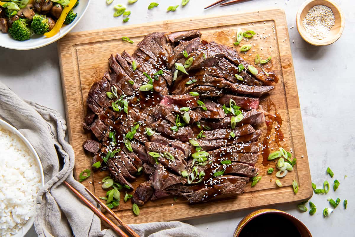 Slices of grilled steak teriyaki on a cutting board surrounded by a bowl of sesame seeds and a bowl of stir fried veggies.