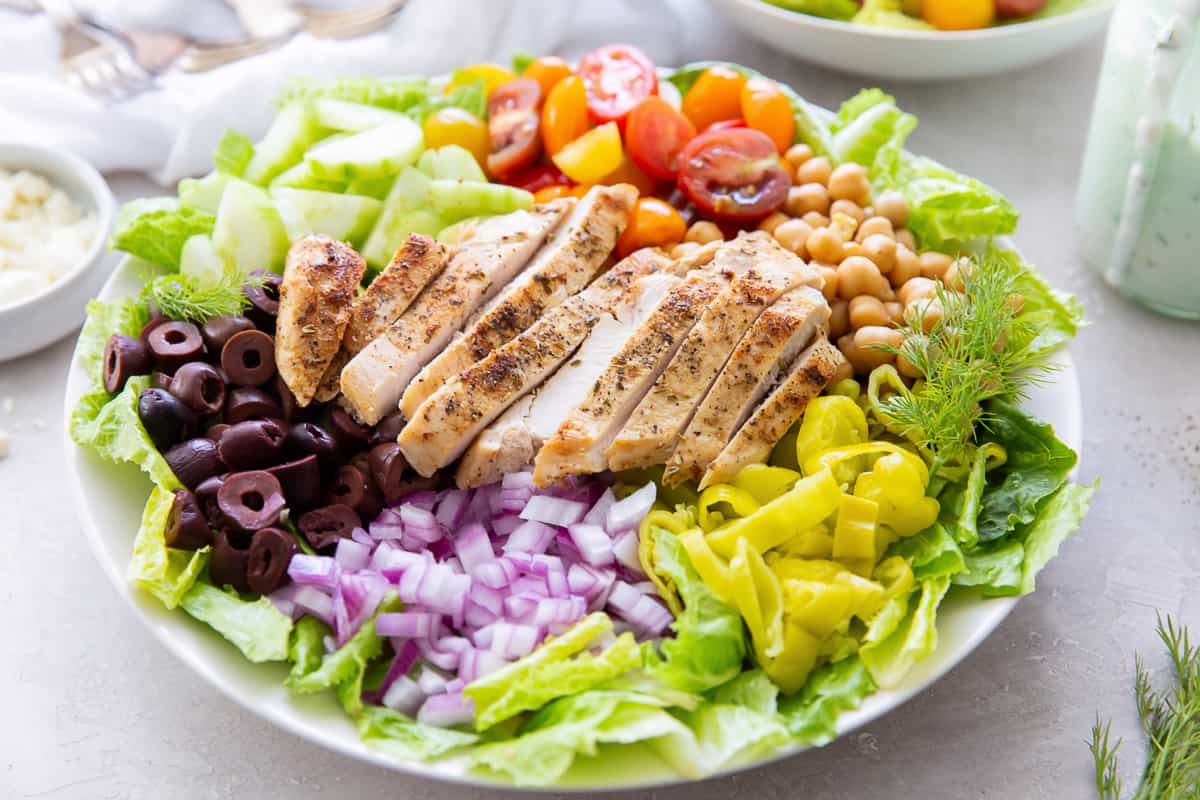 A wide shallow bowl filled with salad topped with chicken, kalamata olives, red onion, and pepperoncini.