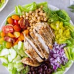 A top down shot of Mediterranean Chicken Salad in a white bowl next to a pepper shaker, feta in a small bowl, and salad dressing.