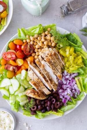 A top down shot of Mediterranean Chicken Salad in a white bowl next to a pepper shaker, feta in a small bowl, and salad dressing.